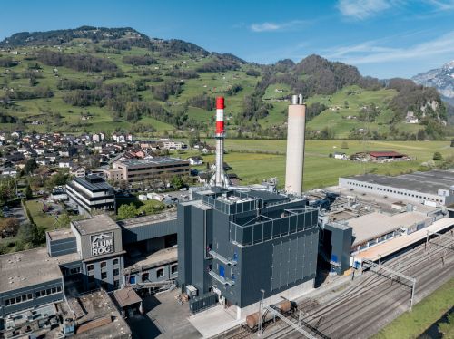 Industrial facility with a red-and-white chimney, surrounded by green hills and a small village under a clear blue sky.