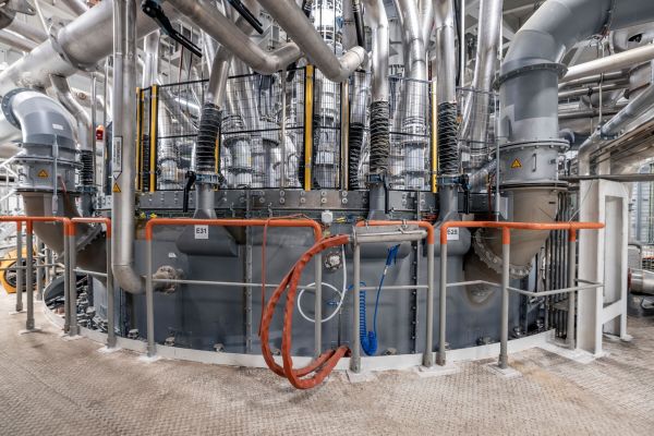 Interior view of industrial machinery with interconnected pipes, cables, and metal components, enclosed by safety barriers.
